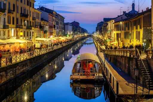 Un canale cittadino scorre tra file di edifici multi-piano. Gli edifici su entrambi i lati sono illuminati da luci calde, con riflessi visibili sull'a sull'acqua scura. Al centro del canale galleggia una barca con copertura chiara e un salvagente arancione. Lungo i lati del canale si trovano camminamenti con ringhiere, caratterizzati da ombrelloni bianchi sulla sinistra e una passerella in legno sulla destra con persone. Il cielo presenta tonalità di blu e pesca.

