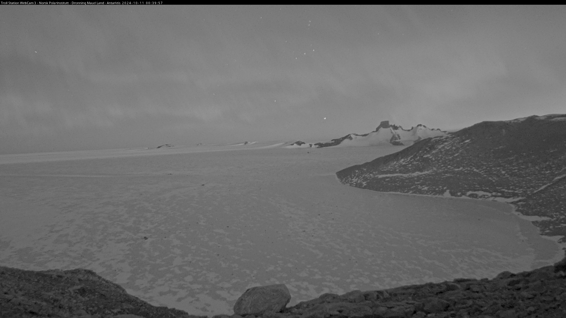 A grayscale nighttime photo taken by a webcam overseeing a large snow-covered plain, with some low rocky hills framing the bottom and the right side of the view, with some more hills visible in the background.

Northern lights are dimly visible in the night sky.