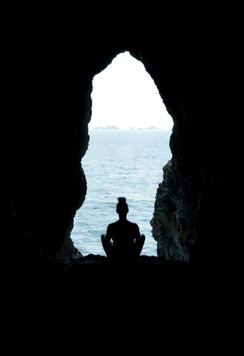 A silhouetted figure sitting in a cross-legged meditation pose inside a large sea cave, facing outward toward the ocean. The cave opening frames the view like a natural window, revealing calm blue ocean waters extending to the horizon where a distant landmass is visible. The cave's interior walls are rough-textured rock that appears dark and shadowy, creating a stark contrast with the bright exterior lighting. 