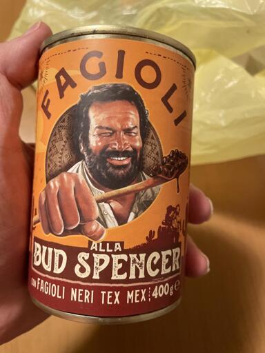 Una mano tiene una lattina metallica cilindrica con un'etichetta dai toni arancione e marrone. L'illustrazione sulla lattina mostra una persona sorridente con barba scura e capelli scuri che impugna un cucchiaio di legno carico di fagioli neri. Il testo sull'etichetta riporta: "FAGIOLI", "ALLA BUD SPENCER", "CON FAGIOLI NERI TEX MEX" e "400ge". Sullo sfondo si vede una borsa di plastica gialla sfocata e una superficie in legno.