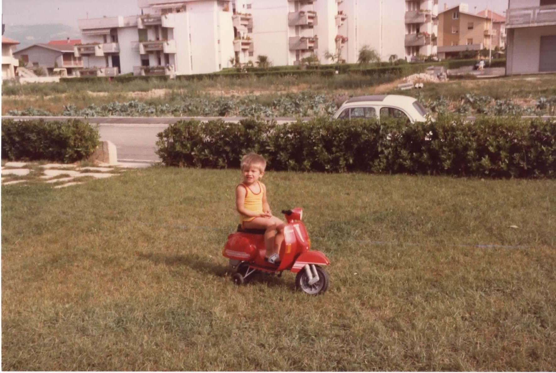 A vintage photograph of a young child (me) seated on a red toy scooter in a grassy area. The child is wearing a yellow tank top and has short blond hair. In the background, multi-story apartment buildings can be seen, as well as a road with a classic Fiat 500 parked nearby. The scene appears to be from about 41 years ago.