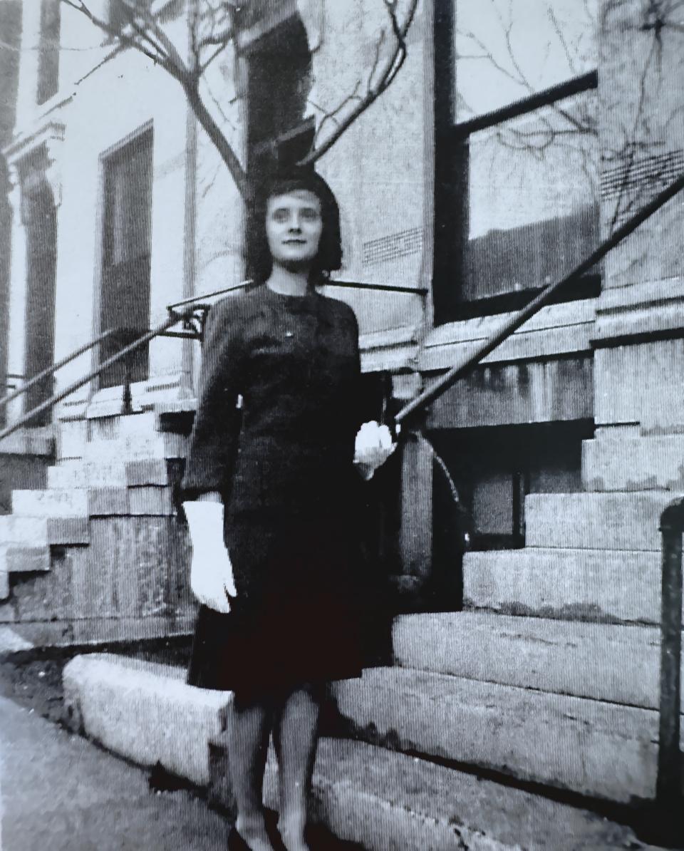 Eva's grandmother during her 20s, having moved to Chicago, standing in front of a residential building's stairway. she gazes upwards to the diagonal-left, holding a jacket while wearing a dress and formal gloves.