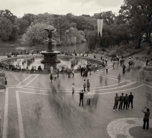 A plaza in a park with a fountain, with a small lake in the background. People, many blurred in motion, mill about.