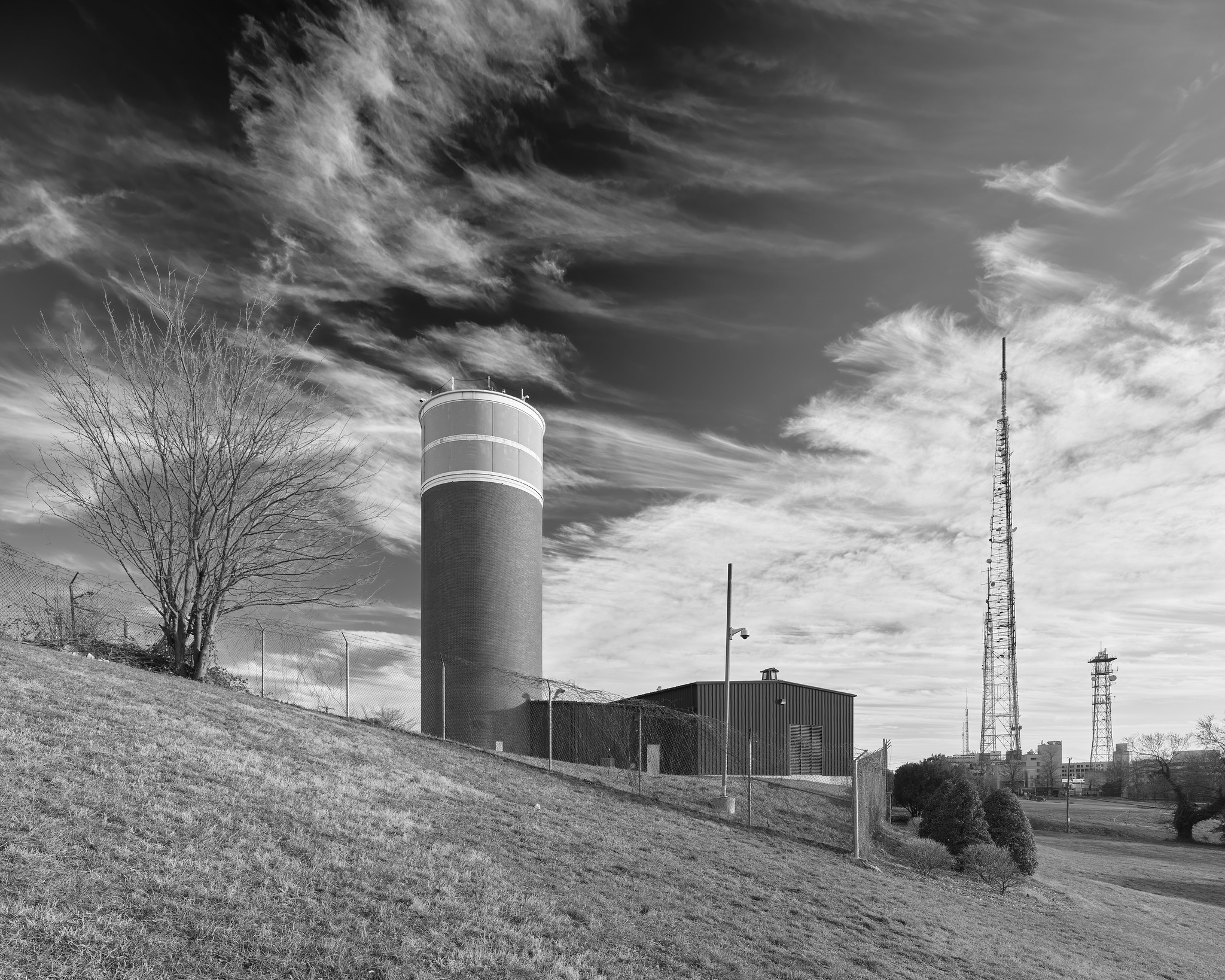 A cylindrical tower, brick with a white top, resembling a water tower or agricultural silo, on a hilltop. Radio towers are visible in the distance. A barren tree is at left.