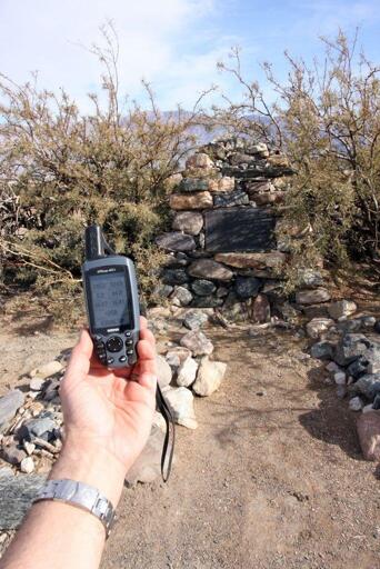 A stone monument set with a bronze plaque has scrub brush growing around it in the desert. There are two sets of stone graves and in the foreground a hand is holding a GPS which shows the elevation at 258 below sea level.