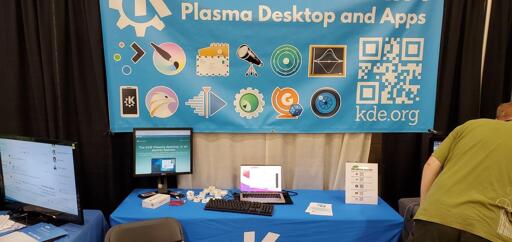 View of KDE's 2023 booth at Scalex20. A banner hangs from the wall, showing a bunch of icons for KDE apps on blue background. Below, the booth itself is loaded with laptops, phones and other devices, as well as stickers. 