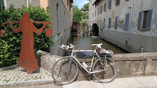 Foto di una bici appoggiata ad un muretto. Dietro ci sono degli edifici in pietra e un canale con un vecchio mulino