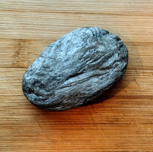 A dark green, shiny, rough oval-shaped stone with visible thin layers, on a wooden cuttin board.