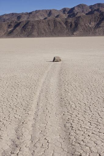 A large rock with a trail behind it sits on the distinctive, cracked playa in Death Valley. Two ridges of brown hills loom up in the background.