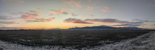 L'immagine mostra un bellissimo paesaggio invernale all'alba scattato dall'aeroporto di Brașov. Le montagne visibili sono parte dei Carpazi, che circondano la città di Brașov, situata in Transilvania.
L'orizzonte è tinto di un arancio intenso e giallo brillante dove il sole sta sorgendo, sfumando gradualmente in toni più tenui di rosa e pesca nelle nuvole sparse sopra. La parte superiore del cielo è di un azzurro-grigio freddo e chiaro.
Una catena montuosa scura e imponente domina il centro dell'inquadratura. Alcune cime sono coperte da un leggero strato di neve, che si staglia contro il cielo illuminato.
Il terreno in primo piano è un campo agricolo spoglio e arato, coperto da un sottile strato di neve bianca, che indica le temperature gelide della stagione invernale.