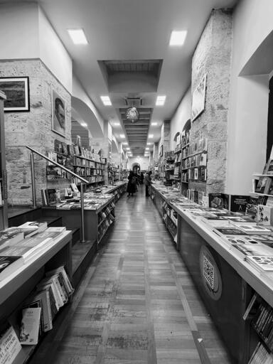 A narrow aisle in a bookstore lined with bookshelves on both sides. The scene is in black and white, featuring wooden flooring and bright overhead lighting. A few people are visible walking in the distance, and images adorn the walls.