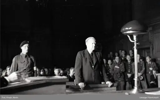 A high contrast black and white photo of a courtroom. A man in the front standing, wearing a suit, a lamp to the right and a soldier to the left. In the background many people are watching