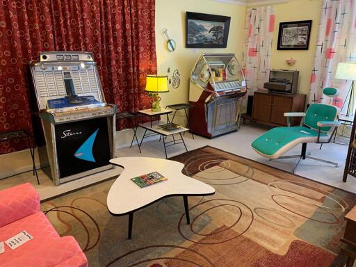 A room filled with retro stylings from the 60s including two jukeboxes, some groovy chairs and tables, a swirly-pattern floor rug, and some wild curtains... YEAH BABY!