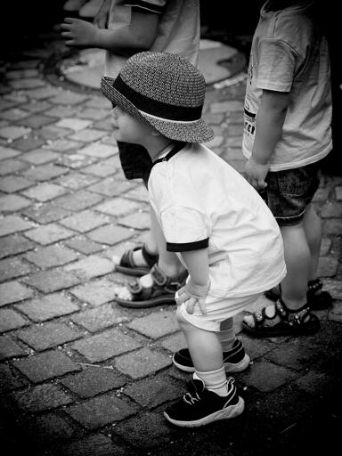 Bambino con cappello di paglia si piega in avanti su un marciapiede di ciottoli, circondato da altri bambini.