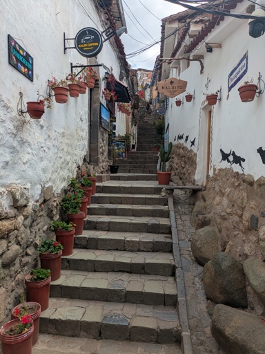 A stair walkway between Incan-era walls composed of stones at the bottom and plaster above, with plants in clay pots along the side and a gutter to direct heavy rainfall. There are shop signs at the top of the stairs saying "Kaldi Coffee Specialty" and "Maldito Never Fucking Surrender 1968"