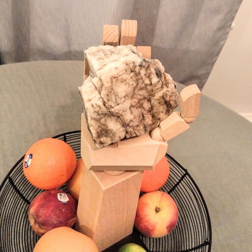 A life-sized wooden hand on a stand holding a baseball-sized white, semi-translucent rock with black veins. The hand rests in a fruit bowl on a table. There are peaches, clementines, asian pears and a lime in the bowl.
