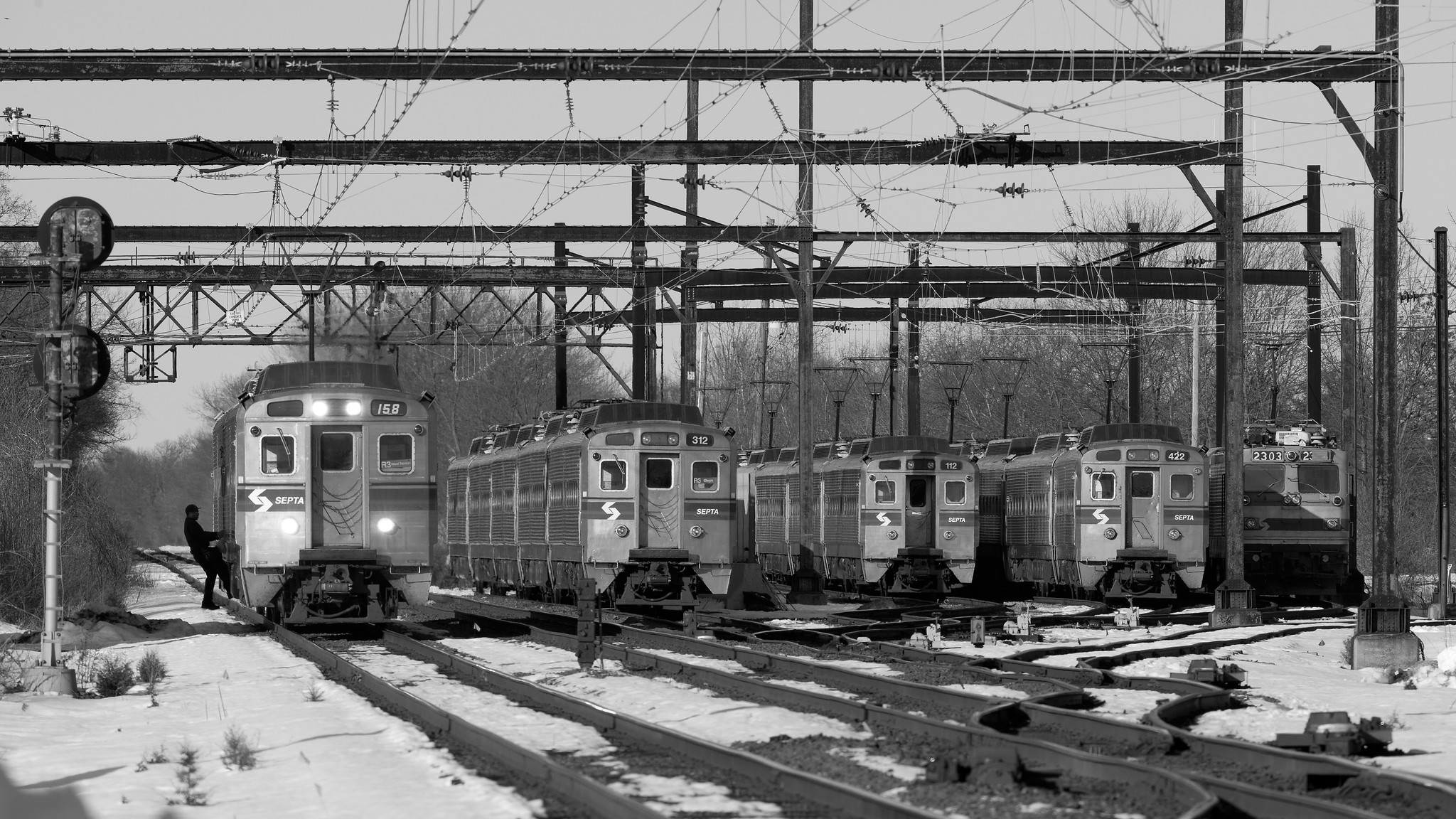 Five electric commuter trains in a rail yard, under overhead wires. A conductor boards the leftmost train. It is winter, with snow on the ground.