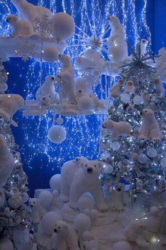Questo immagine mostra una mostra festiva di inverno con numerosi cubi polari bianchi (sia adulti che cuccioli) disposti sui scaffali e intorno a due alberi di Natale decorati. Gli alberi sono adornati con ornamenti bianchi e azzurro chiaro, decorazioni a neve, luci a stella bianche e luci di fili scintillanti. Aggiunte alla mostra sono ulteriori ornamenti cubici bianchi e decorazioni a forma di snowflake, insieme a luci che creano un effetto stellato contro uno sfondo blu profondo. La scena è piena di giocattoli di polar bear in peluche di varie dimensioni, posizionati tra i decorazioni innevate e gli alberi. Non è visibile alcun testo nell'immagine.