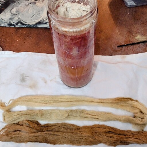 Three strips of wool on towel, died brown/gray ar the bottom, peach in the middle, light peach on top. A tall glass jar of bubbles sits above. 