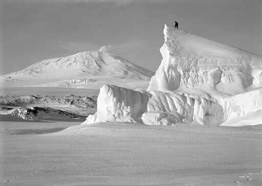 Antarctic explorer stands atop an iceberg, with active volcano Mt. Erebus in the distance, 1911