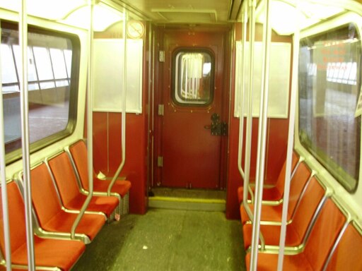 The interior of a small rapid transit train. The lighting is florescent and yellow. The train is small with standing room for two across between the rows of seats which line the car facing inwards. 