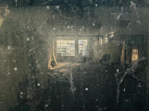 An image of a dusty, dimly lit interior space, possibly a workshop or old building, with large windows letting in light. The view is obscured by grime or reflections, creating a moody atmosphere.