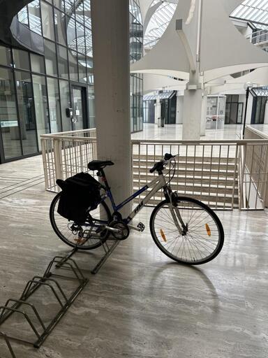 A bicycle is parked on a bike rack inside a modern, spacious building with large glass walls. The interior features marble flooring and a distinctive ceiling design.